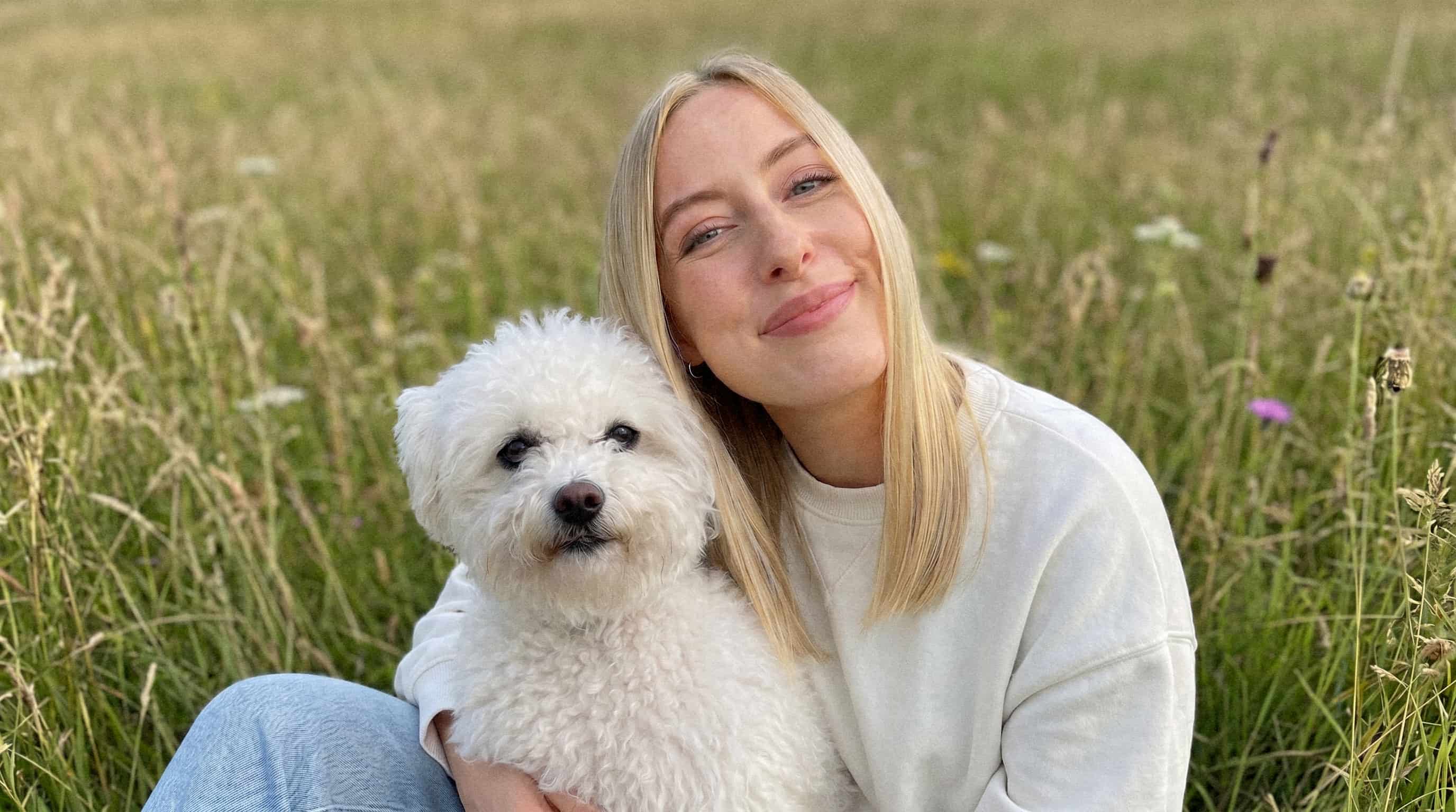 Frau mit blondem Haar sitzt im Gras und lächelt, hält einen weißen Hund mit lockigem Fell.