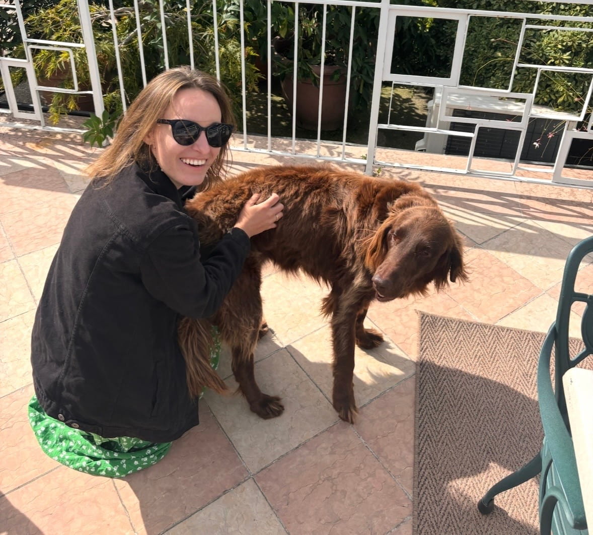 Frau mit langen Haaren und Sonnenbrille kniet neben einem braunen Hund auf einer Terrasse.