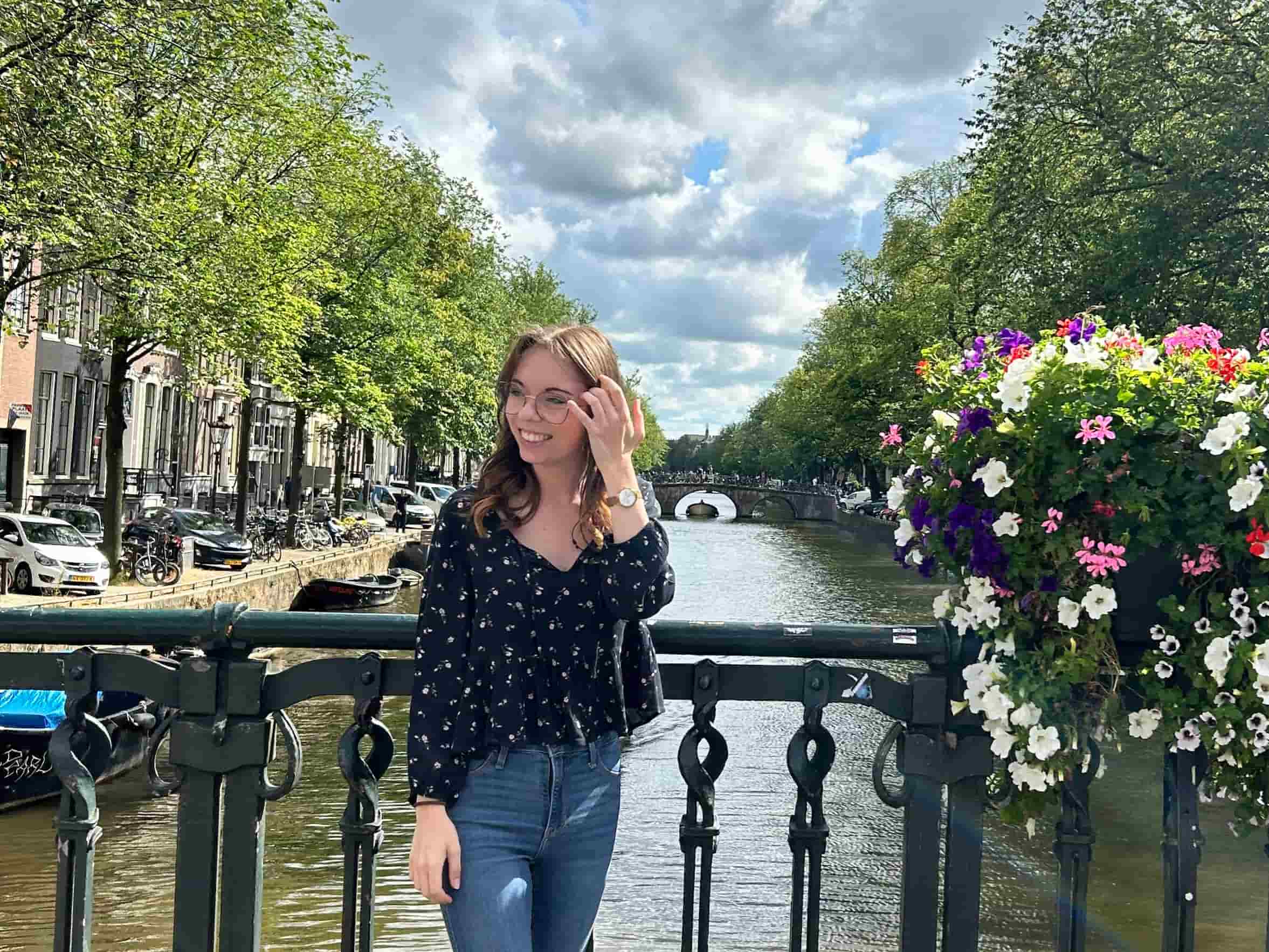 Junge Frau mit langen Haaren, die in einem blütenverzierten Top an einer Brücke in Amsterdam posiert.