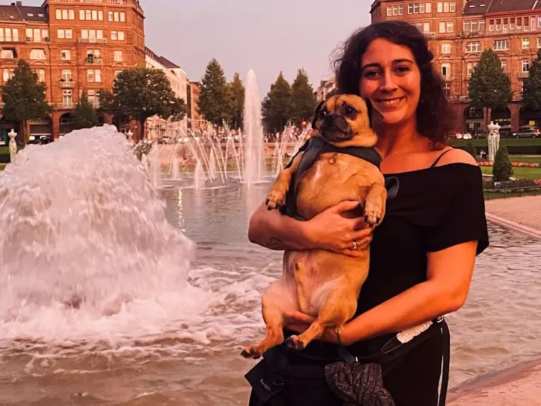 Frau mit Hund vor einem Brunnen in einem Park, umgeben von Bäumen und Gebäuden bei Sonnenuntergang.