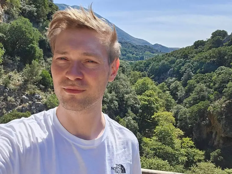 Mann in der Natur mit Blick auf bewaldete Hügel und Sonne, trägt ein weißes T-Shirt der Marke The North Face.