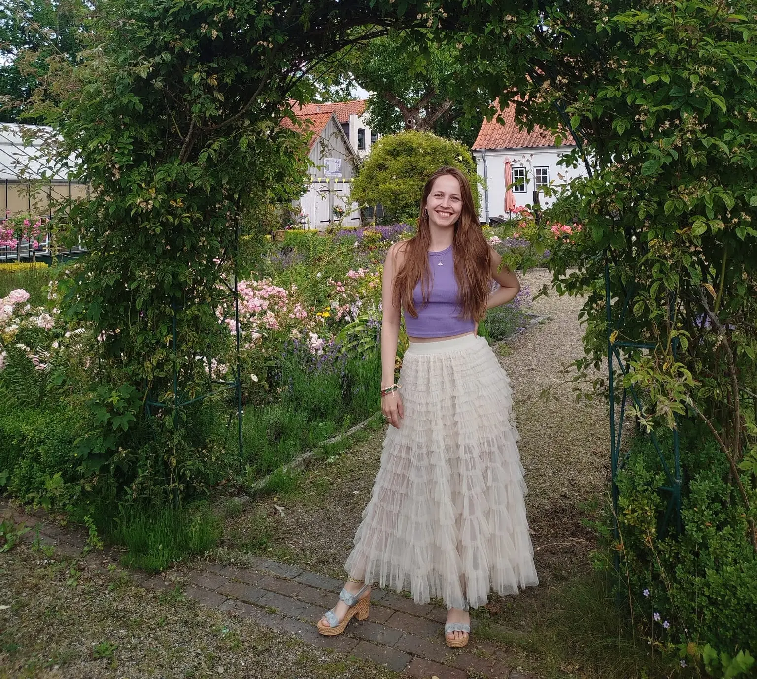 Frau in einem Garten, trägt einen langen, luftigen Rock und ein Top, steht unter einem Bogen aus Pflanzen.