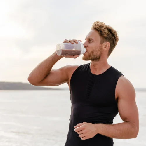 Mann in schwarzem Muskelshirt trinkt einen Shake am Strand mit Meerblick.