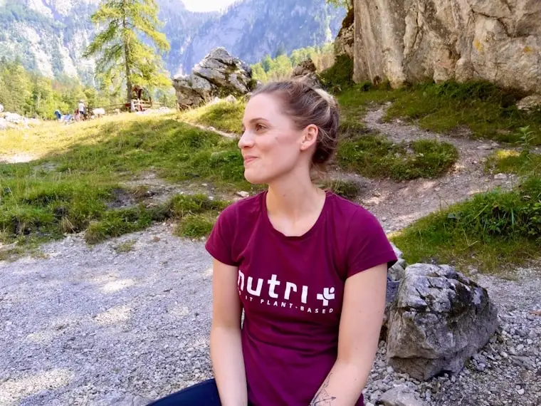 Frau in lila nutri T-Shirt, sitzt auf dem Boden in einer bergigen Landschaft mit Bäumen.