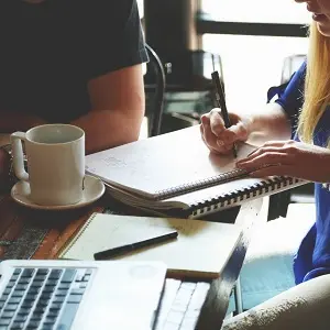 Personen bei einer Besprechung in einem Café mit Notizbuch, Laptop und Kaffeetasse auf dem Tisch.