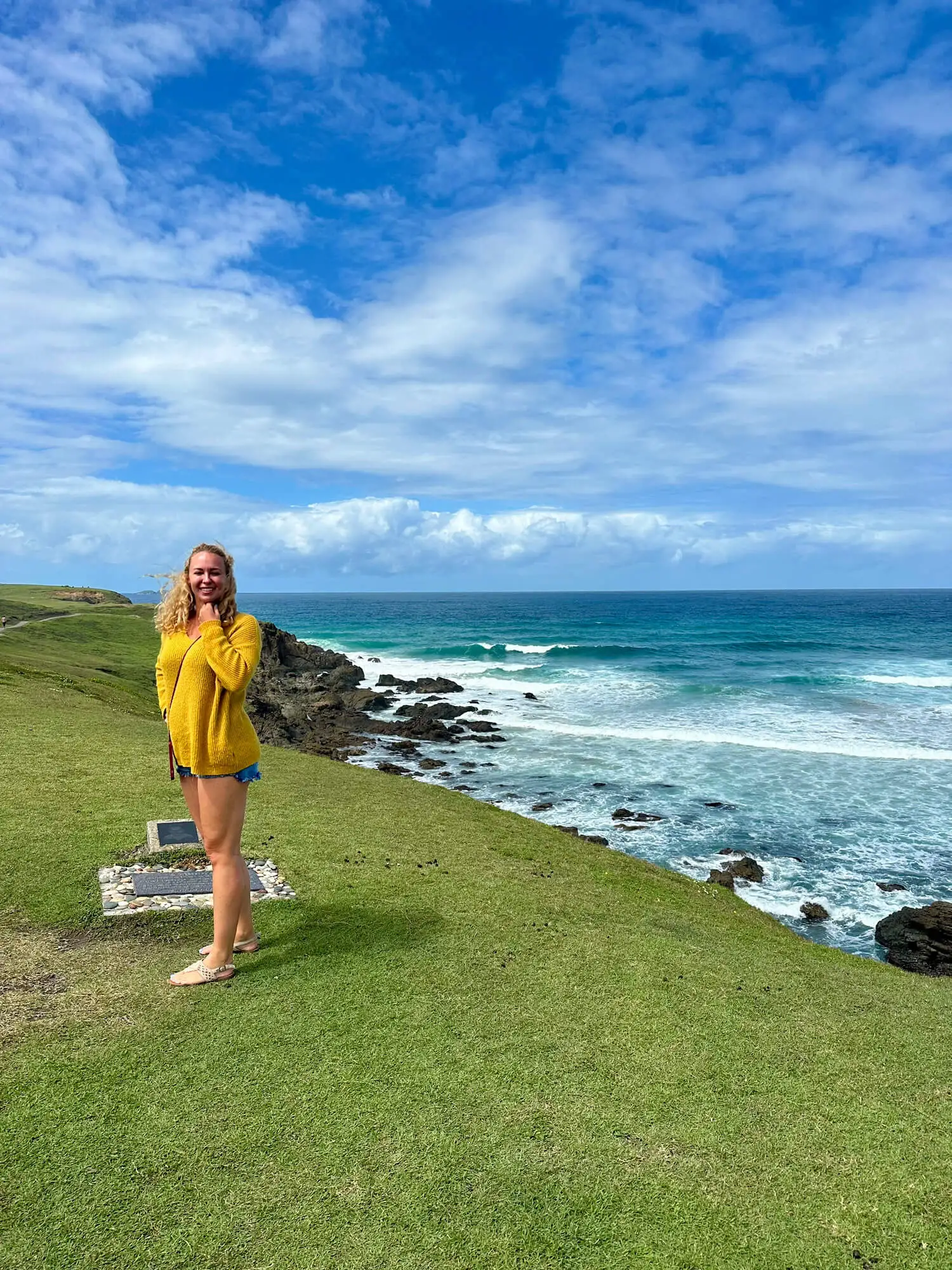 Frau in gelbem Pullover steht am Strand mit Blick auf das Meer und blauen Himmel.